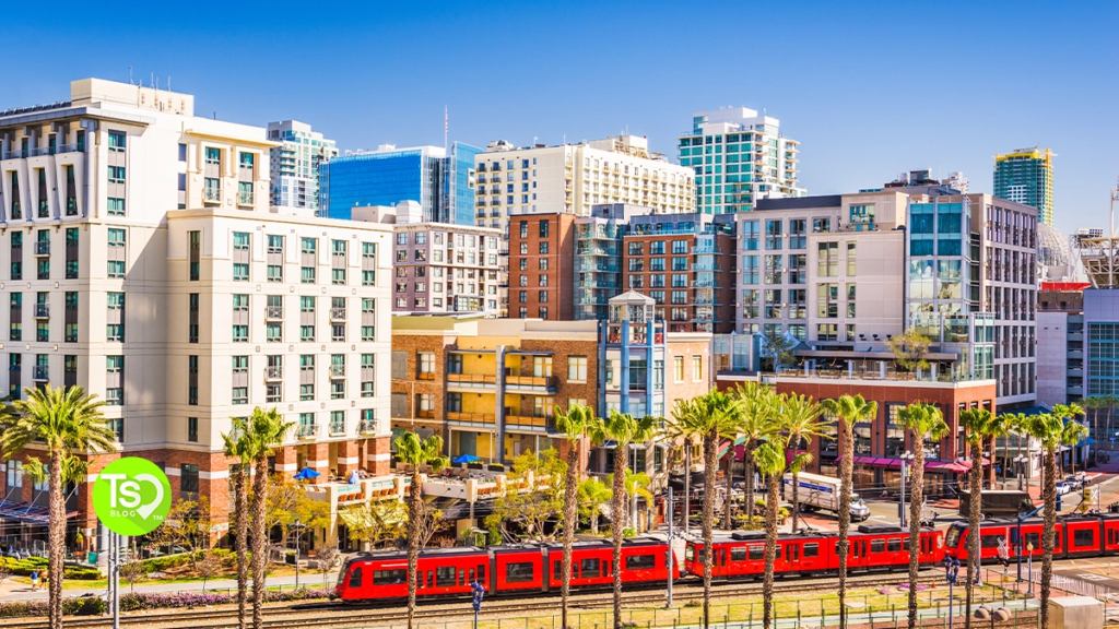 A bright red passenger trolley passing by modern downtown buildings, hotels, and palm trees in sunny San Diego.