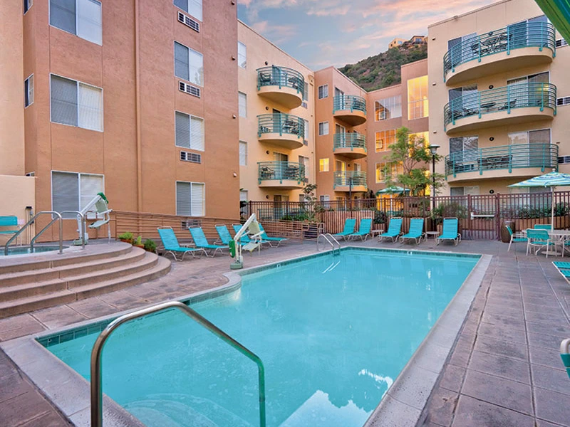 Outdoor swimming pool, hot tub, and bright teal lounge chairs surrounded by the multi-story exterior of the San Diego Mission Valley resort.