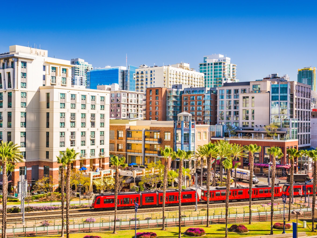 A bright red passenger trolley passing by modern downtown buildings, hotels, and palm trees in sunny San Diego.