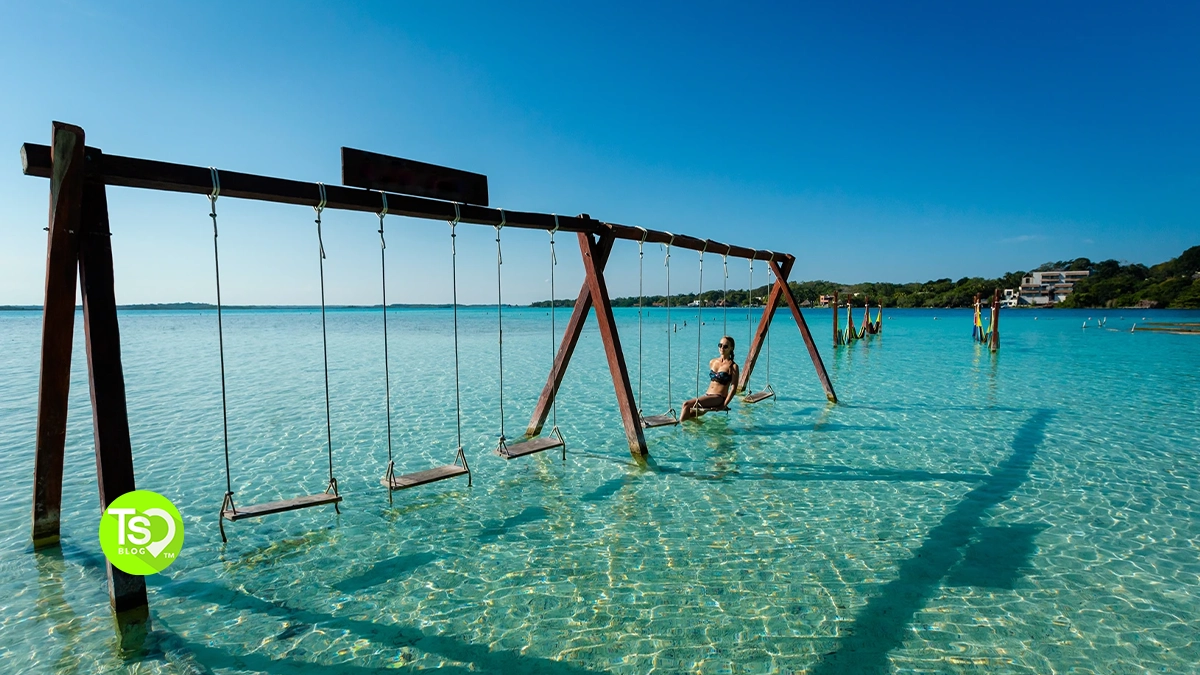 A woman swings over clear turquoise water with a blue sky in the background.