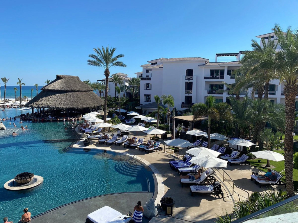 A sprawling resort swimming pool with a large thatched-roof palapa bar, lounge chairs, and palm trees.
