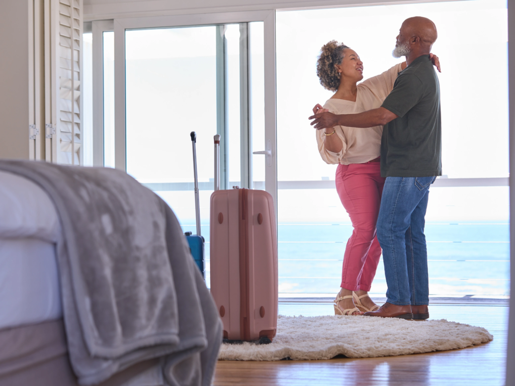 A senior couple happily dancing in a modern hotel room with suitcases near windows overlooking the ocean.