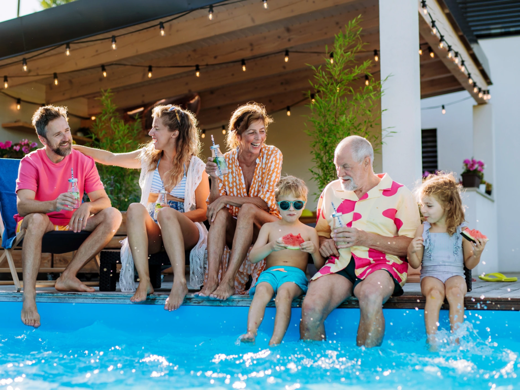 A multi-generational family sitting on a pool deck, laughing, drinking, and eating watermelon slices.