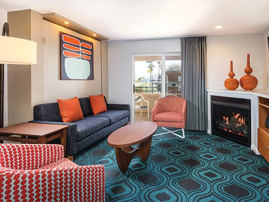A modern resort living room with a geometric teal rug, a dark sofa, coral accent chairs, and a fireplace.