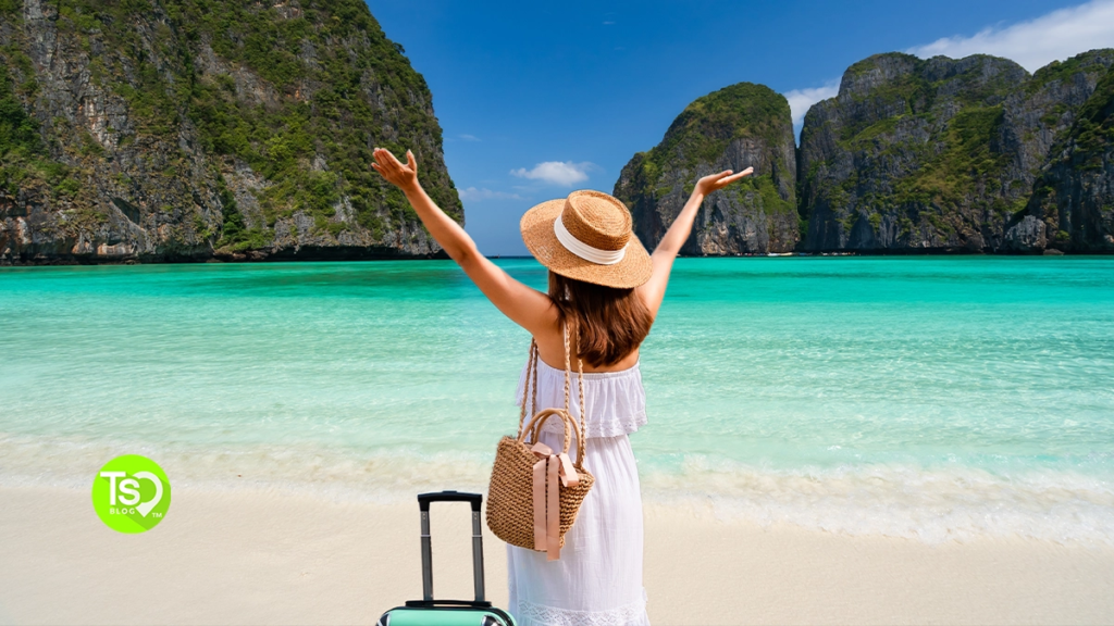 A woman in a white dress and sun hat stands on a white sand beach with her arms raised, perfect for warm weekend getaways.
