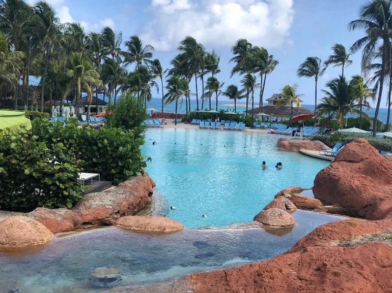 Large tropical swimming pool surrounded by palm trees, red rock formations, and lounge chairs under a cloudy sky.