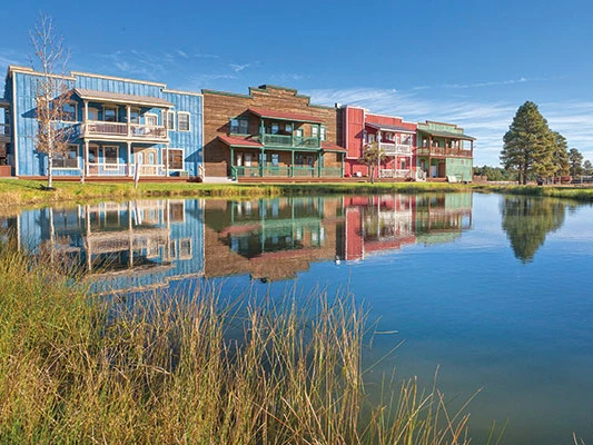 Wild West-style buildings reflecting in a serene pond at Club Wyndham Bison Ranch under a clear blue sky.