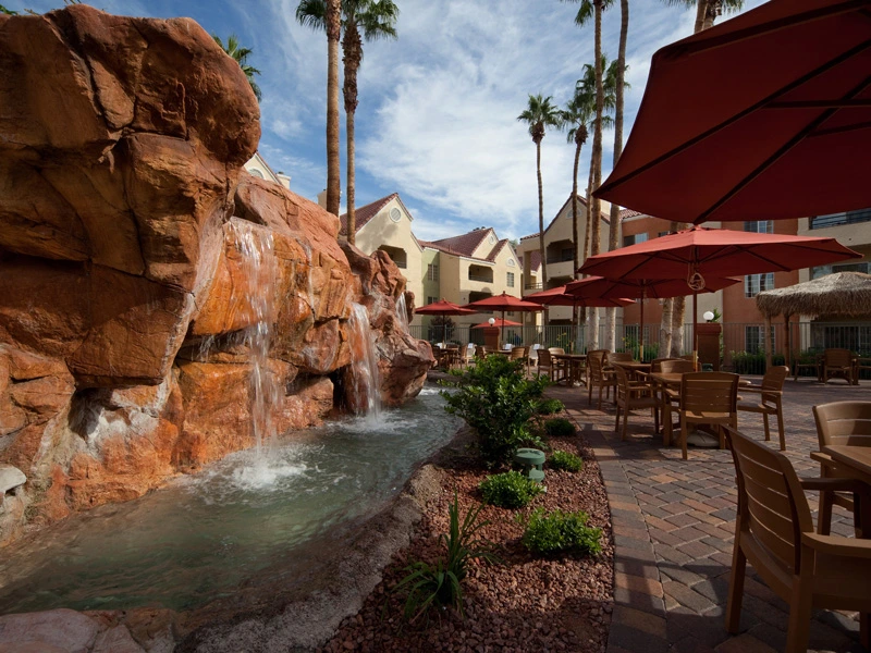 Outdoor patio, red umbrellas, and a large red rock waterfall feature pouring into a pool at Desert Club Resort.