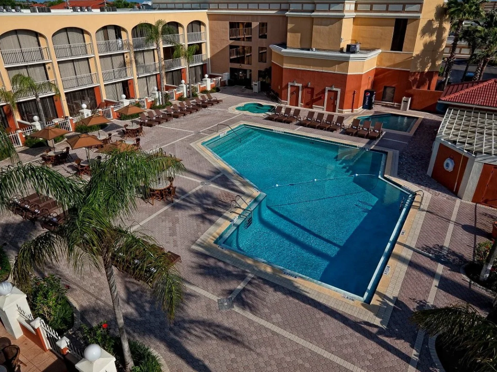 Aerial view of a hotel courtyard with a large outdoor pool, lounge chairs, umbrellas, and palm trees.