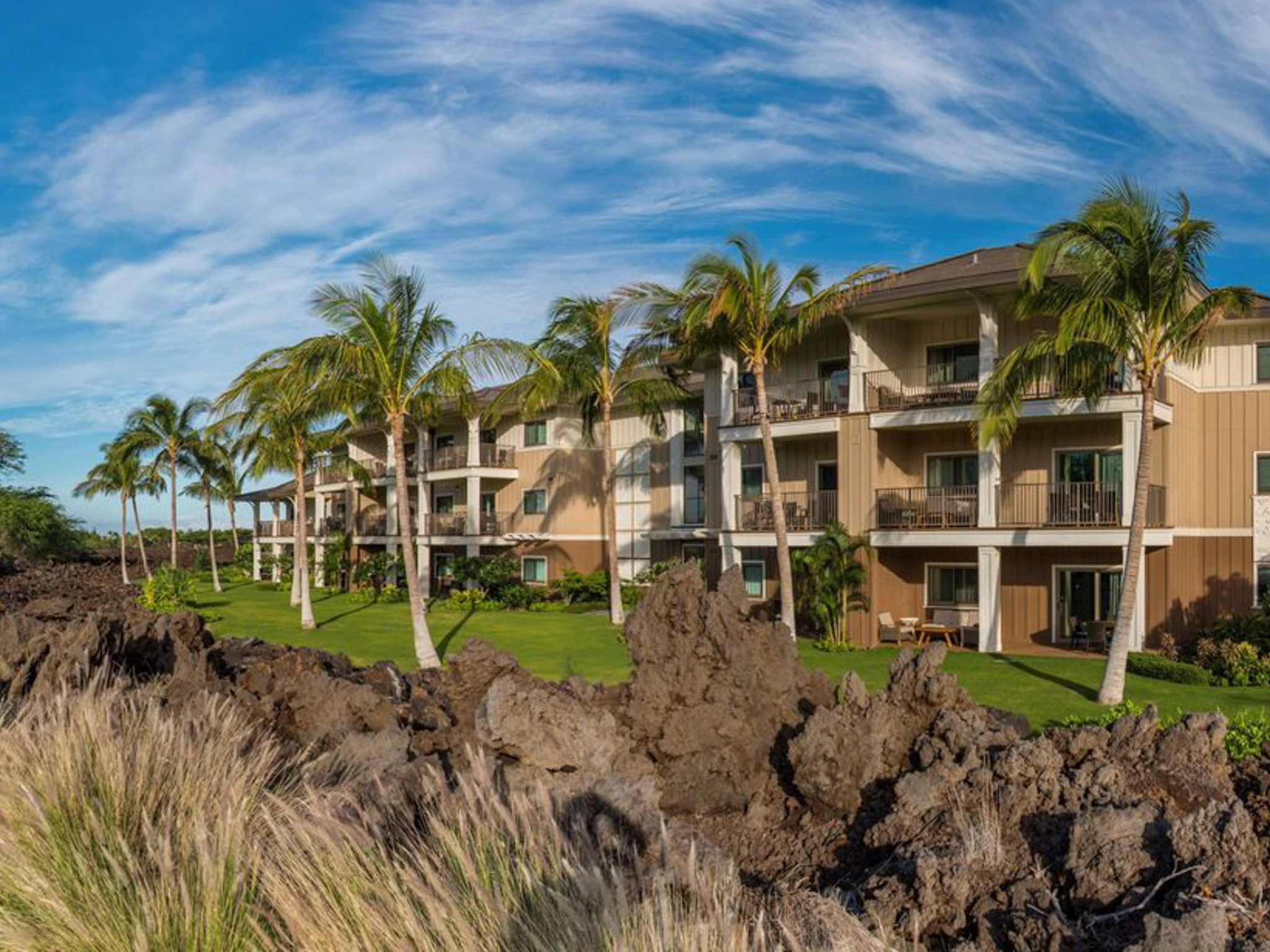 Exterior of Kings' Land resort featuring a green lawn, palm trees, and lava rocks.
