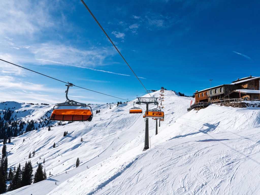 Orange chairlifts carry skiers up a snow-covered mountain slope beneath a clear blue sky.