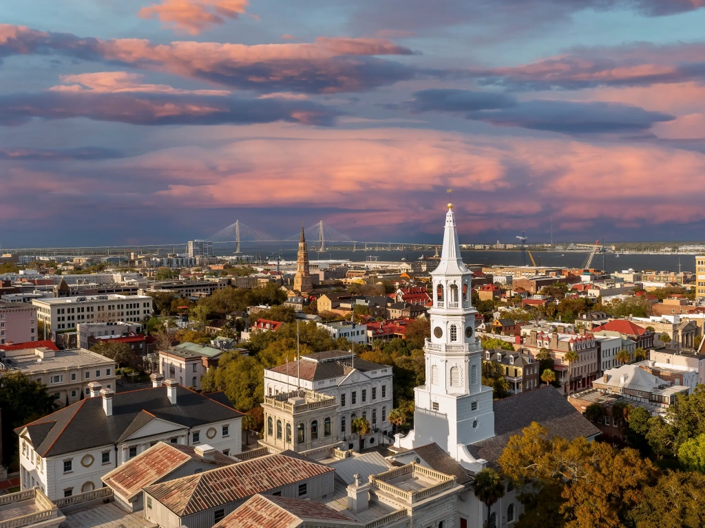 Aerial view of Charleston South Carolina skyline at sunset with a white church steeple and harbor bridge in background.