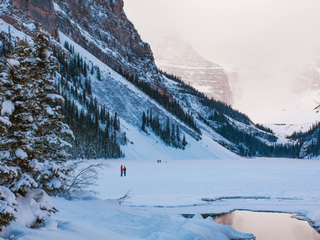 Two hikers in colorful jackets explore a frozen mountain lake, perfect for Romantic Getaways amid snowy cliffs and evergreen trees.