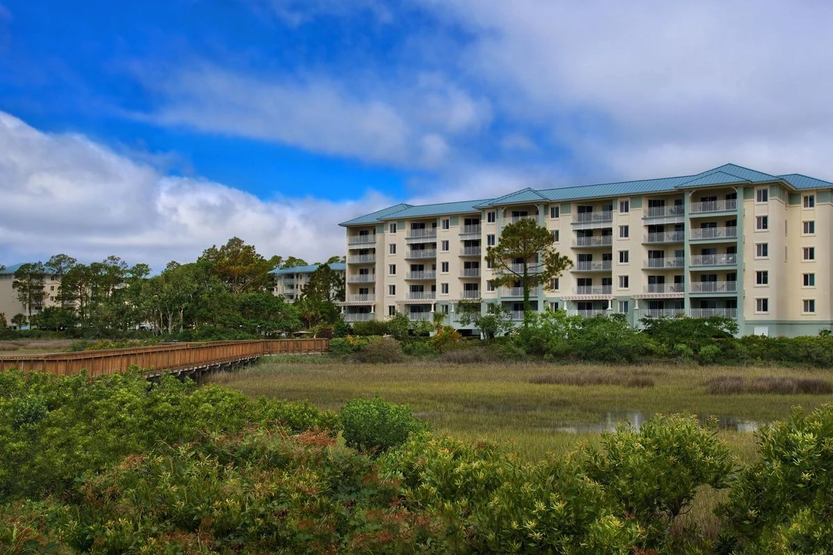 A multi-story coastal resort building viewed across a lush green marshland and a wooden boardwalk under a blue sky.