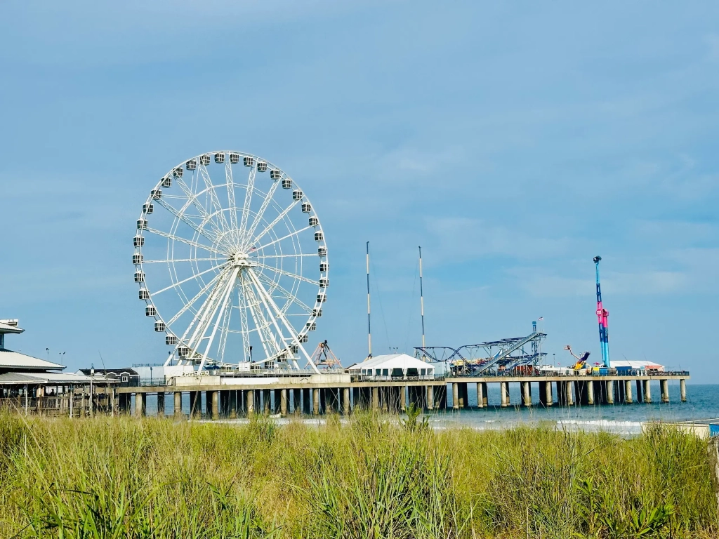 A Ferris wheel rises over one of the best boardwalks on east coast, with an amusement pier, ocean waves, and dune grass below.