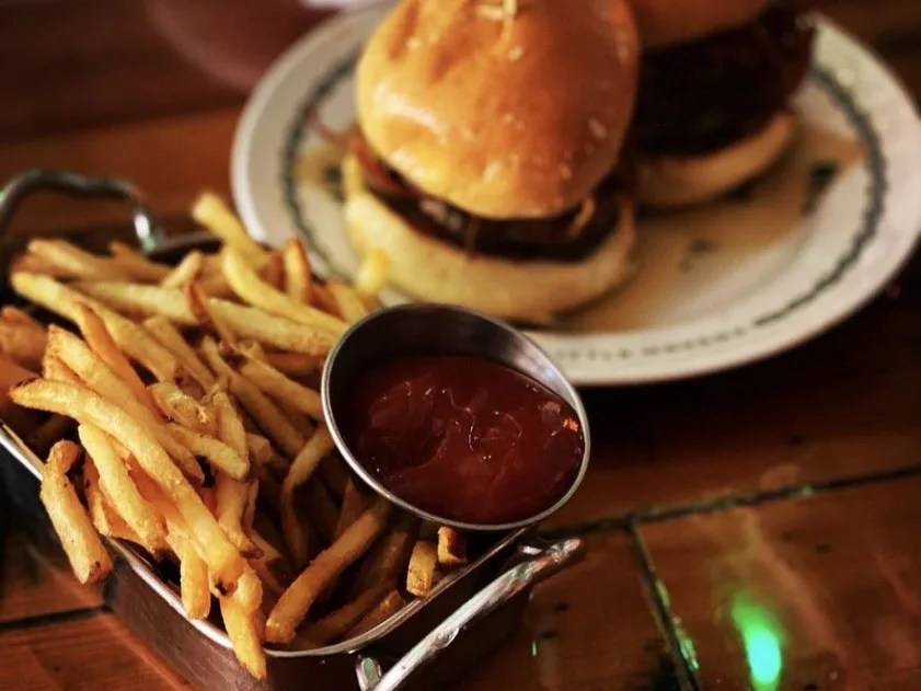 Crispy french fries in a metal basket with ketchup dipping cup beside a burger on a wooden table.