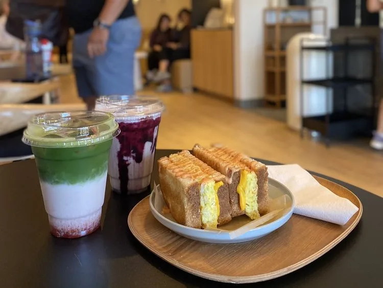 Layered matcha latte, berry drink, and a toasted egg sandwich cut in half on a wooden tray at a cafe