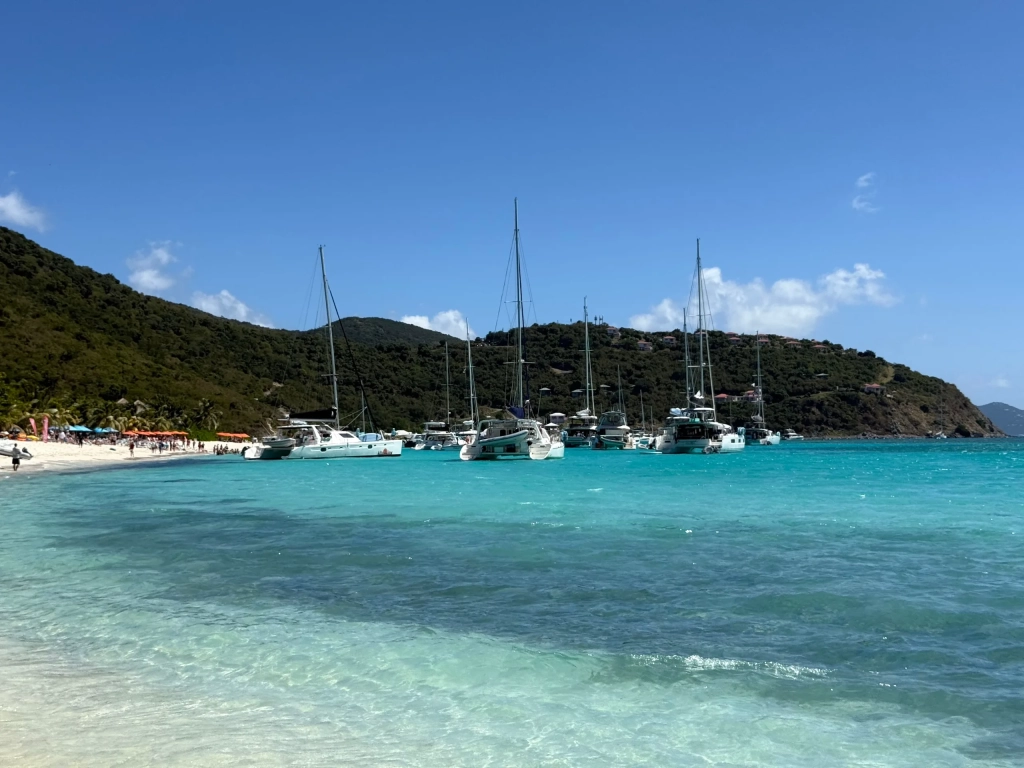 Sailboats anchored in turquoise Caribbean waters, perfect for festiva sailing vacations, near a white sand beach backed by green hills.