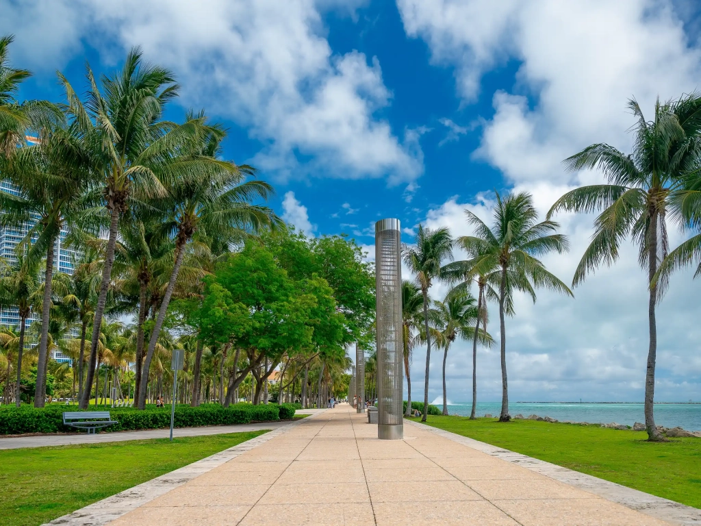 Palm-lined waterfront promenade with a tall metal sculpture, green lawn, and blue sky along a coastal park walkway.