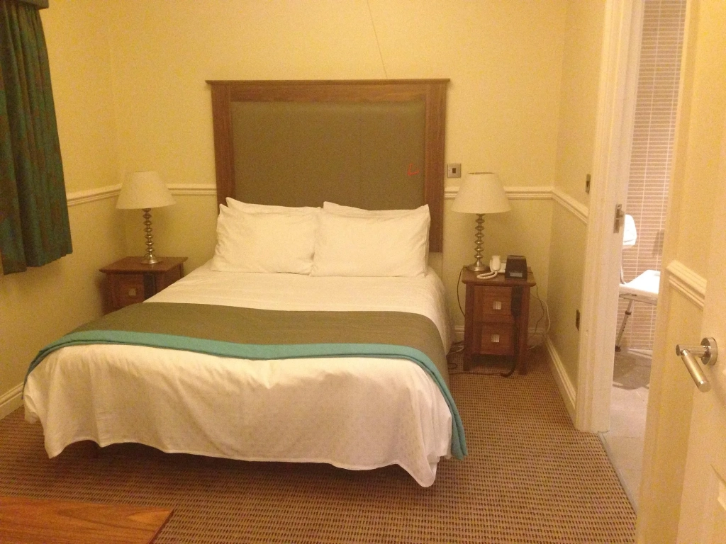 Double bed with white linens, a teal throw, and a green headboard between two bedside lamps, with an open door to the en suite bathroom.
