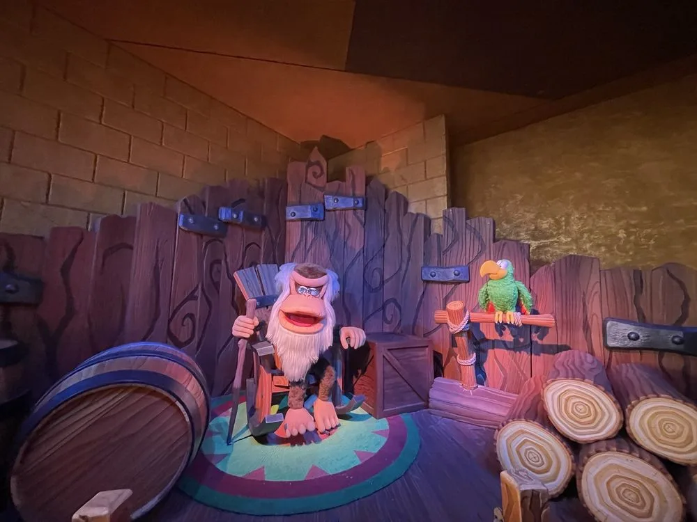 Animatronic ape character with white beard and glasses sits beside a green parrot in a rustic wooden themed attraction scene.