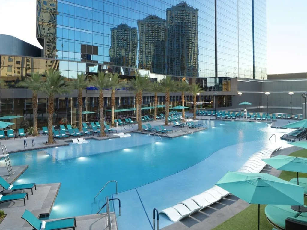 Large outdoor resort pool surrounded by teal lounge chairs and palm trees with mirrored glass towers reflecting in the background.