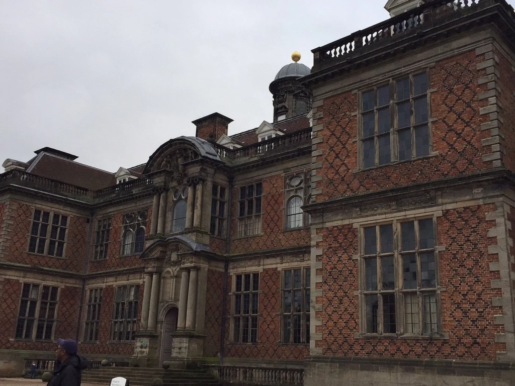 A grand English Baroque country house with ornate red brick diaper patterning, stone pilasters, and a central pediment above the entrance.