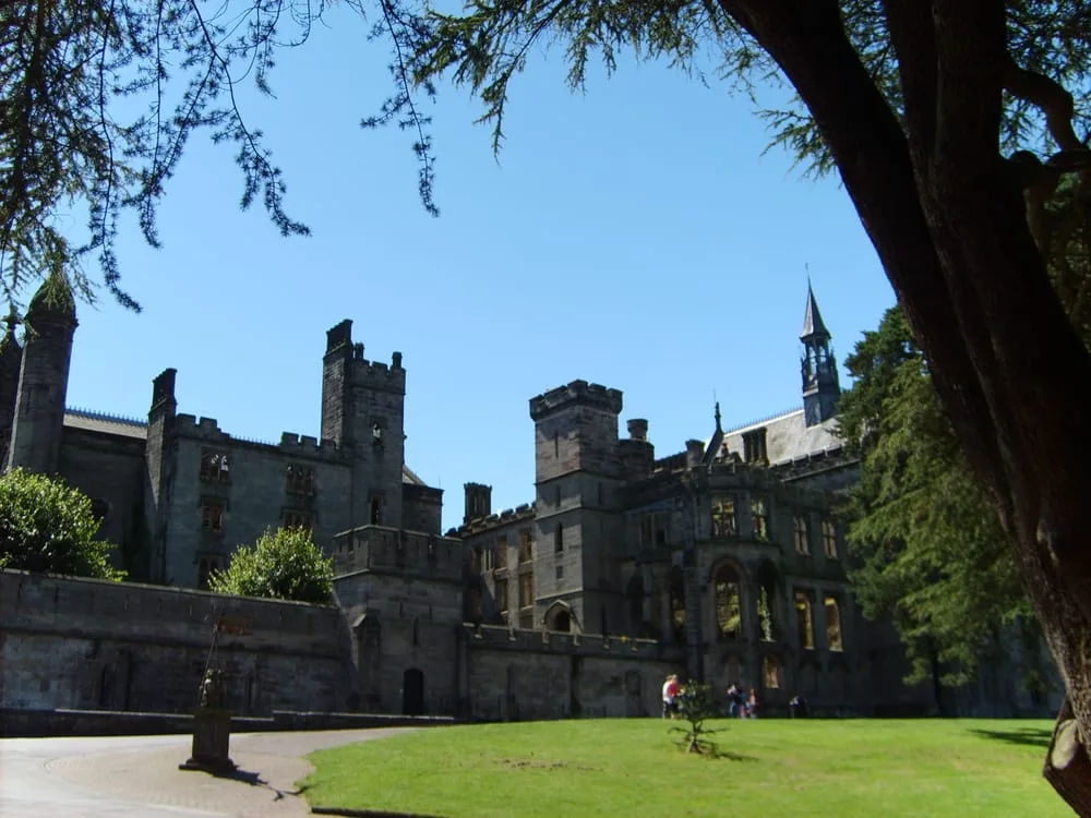 Gothic revival castle with battlements and a pointed spire surrounded by green lawns and mature trees on a sunny day.