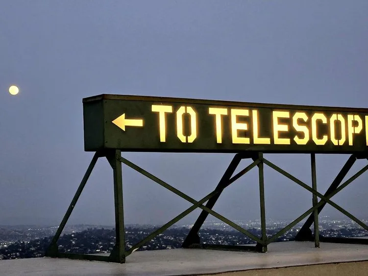 Illuminated sign reading To Telescope with an arrow pointing left at Griffith Observatory at dusk with moon and city lights
