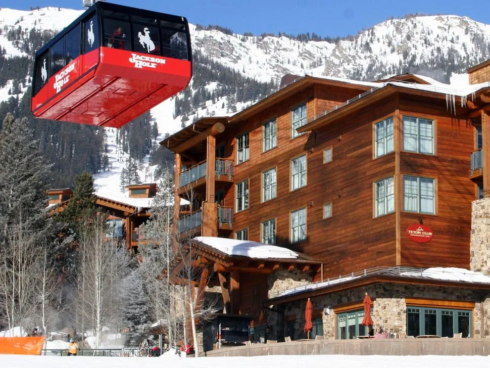 Red Jackson Hole aerial tram gondola passes above a snow-covered rustic wood and stone lodge at the base of a mountain.