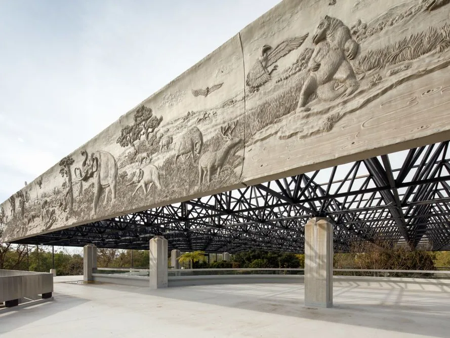 Large concrete beam with detailed bas-relief carvings of animals and wildlife above a steel lattice canopy structure.