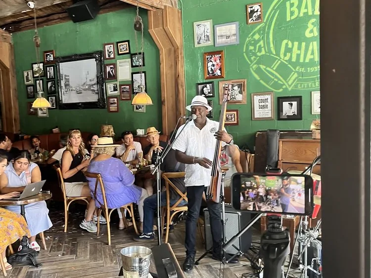 A musician in a white hat plays upright bass and sings at a mic in a green-walled jazz bar.