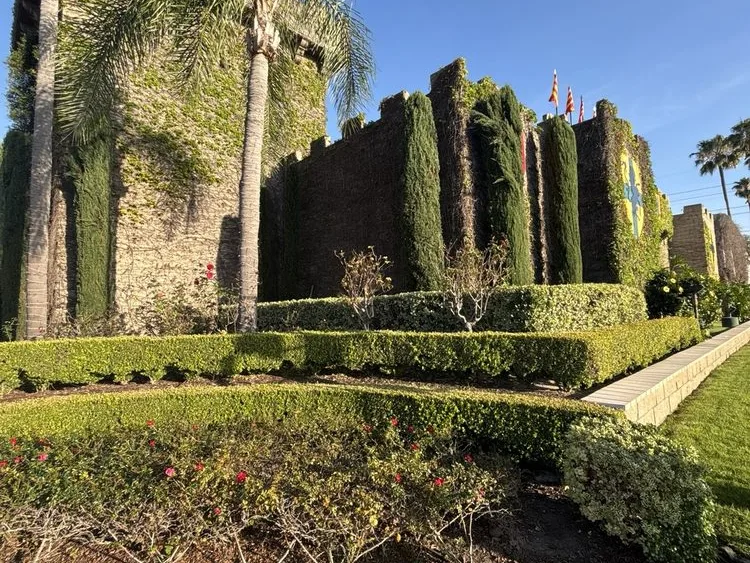 Castle-style stone walls draped in ivy with manicured hedges, palm trees, and colorful flags under a clear blue sky.