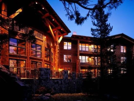 Rustic wood and stone lodge exterior glowing with warm interior lights against a twilight sky surrounded by pine trees.