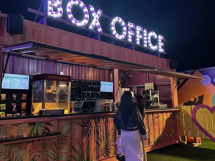 A woman explores Things To Do in Miami At Night at a glowing outdoor Box Office concession stand with popcorn and a menu board.