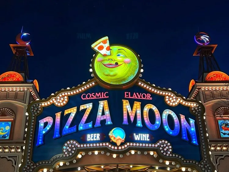 Neon Pizza Moon restaurant sign near Epic Universe tickets booth, showing a smiling moon mascot holding a pizza slice.