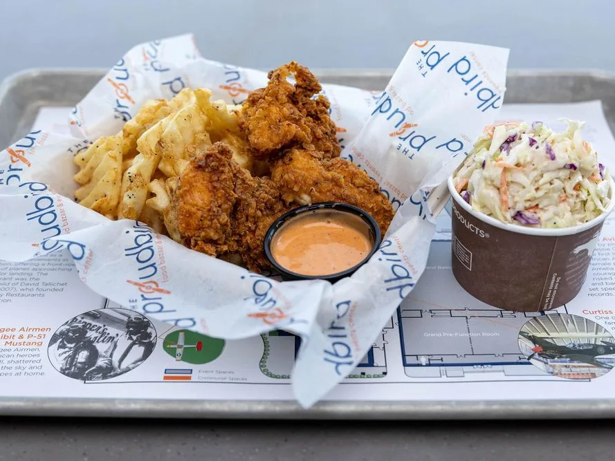 Crispy fried chicken tenders with seasoned fries, dipping sauce, and coleslaw on a branded tray liner.