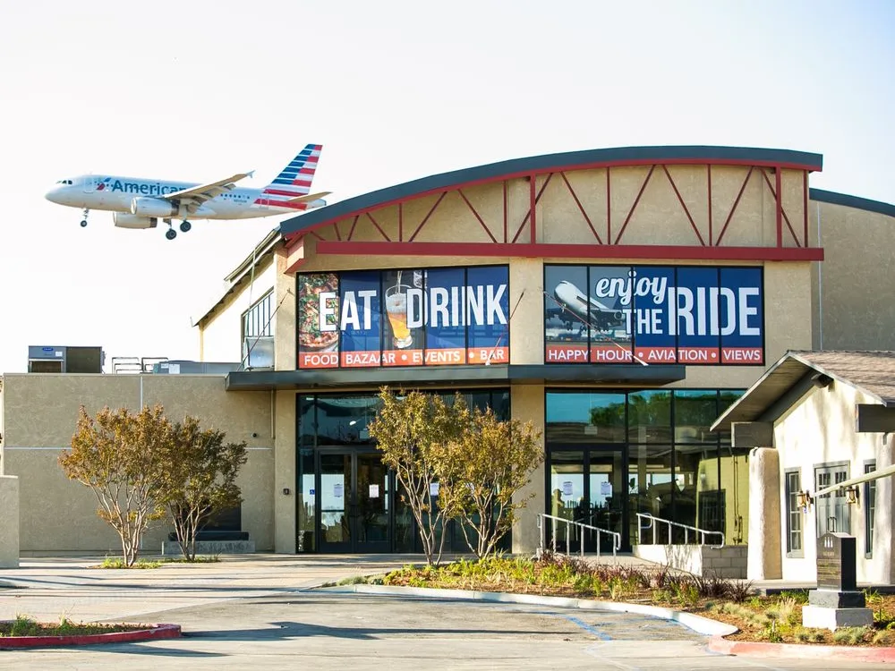 American Airlines jet flying low over the Proud Bird restaurant near Los Angeles Airport, with Eat Drink and Enjoy the Ride signs visible.