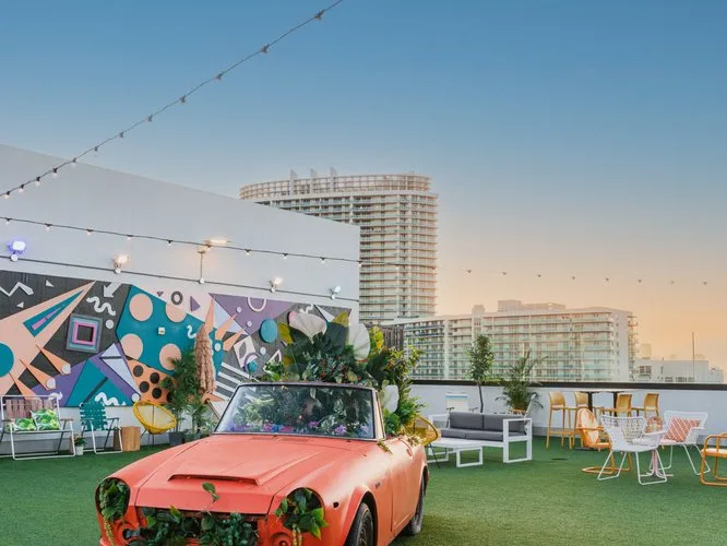Coral pink vintage convertible car filled with plants on a rooftop terrace with colorful geometric mural and city skyline.