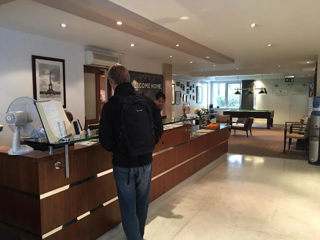 Traveler with backpack checking in at a hotel front desk with a Welcome Home sign and billiard table in background.