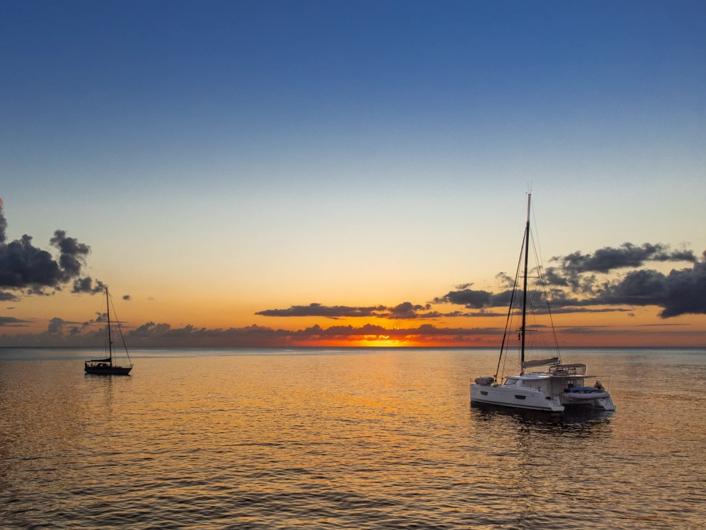 Two sailboats anchored on calm golden water at sunset with a vivid orange horizon and scattered clouds.