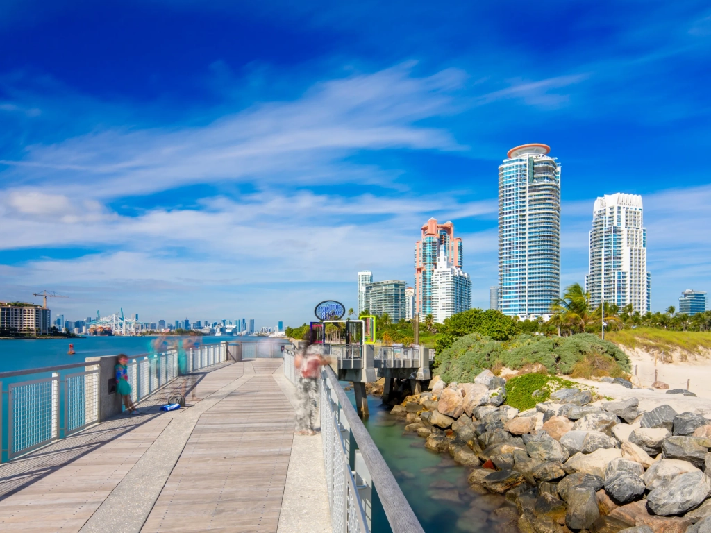 Wooden pier extending along the waterfront in South Beach Miami with luxury high-rise condos and blue sky.