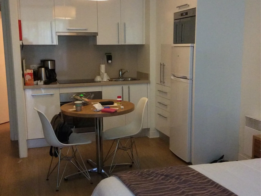 Compact studio apartment with white kitchen cabinets, small round dining table, two white chairs, and a bed in the foreground.