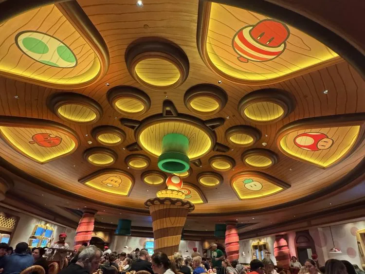 Crowded dining room at Epic Universe tickets venue with Mario mushrooms, glowing panels, and a green pipe ceiling centerpiece.