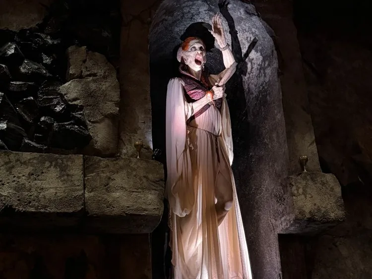 A vampire character in dramatic makeup and flowing white gown poses in a stone dungeon setting.