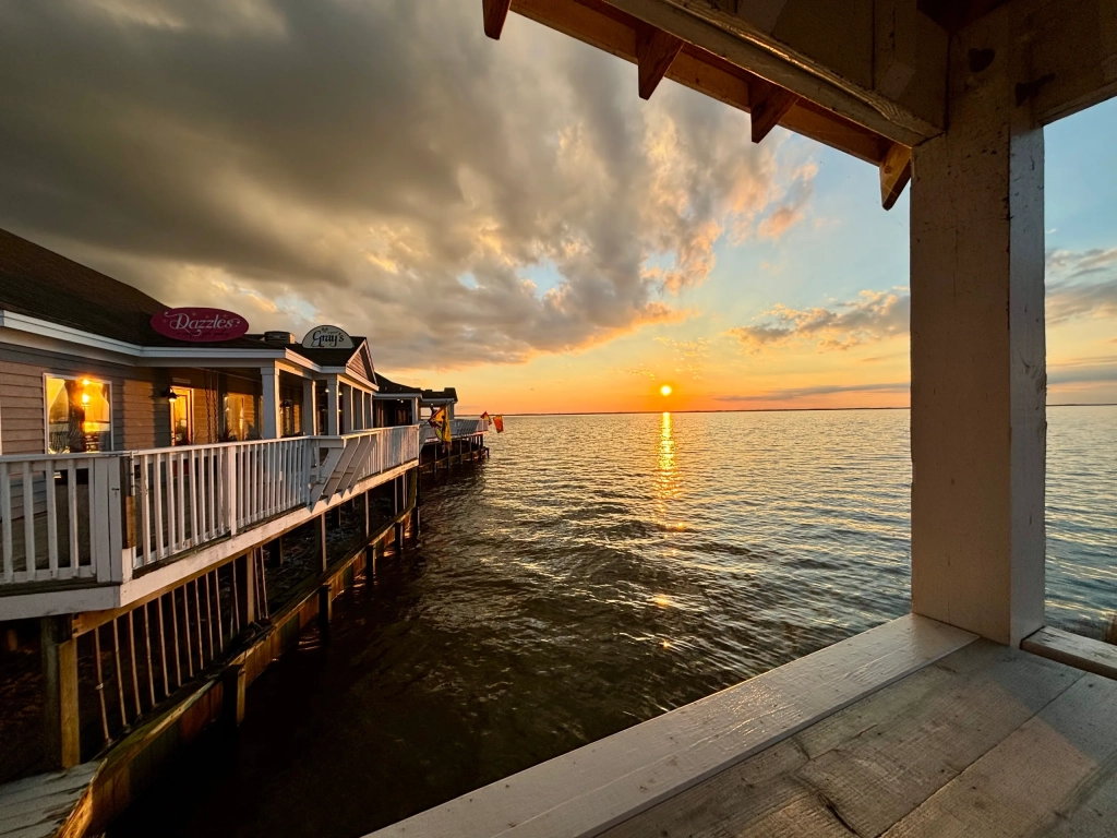 Waterfront restaurants on a pier extending over calm water at sunset with dramatic clouds overhead.