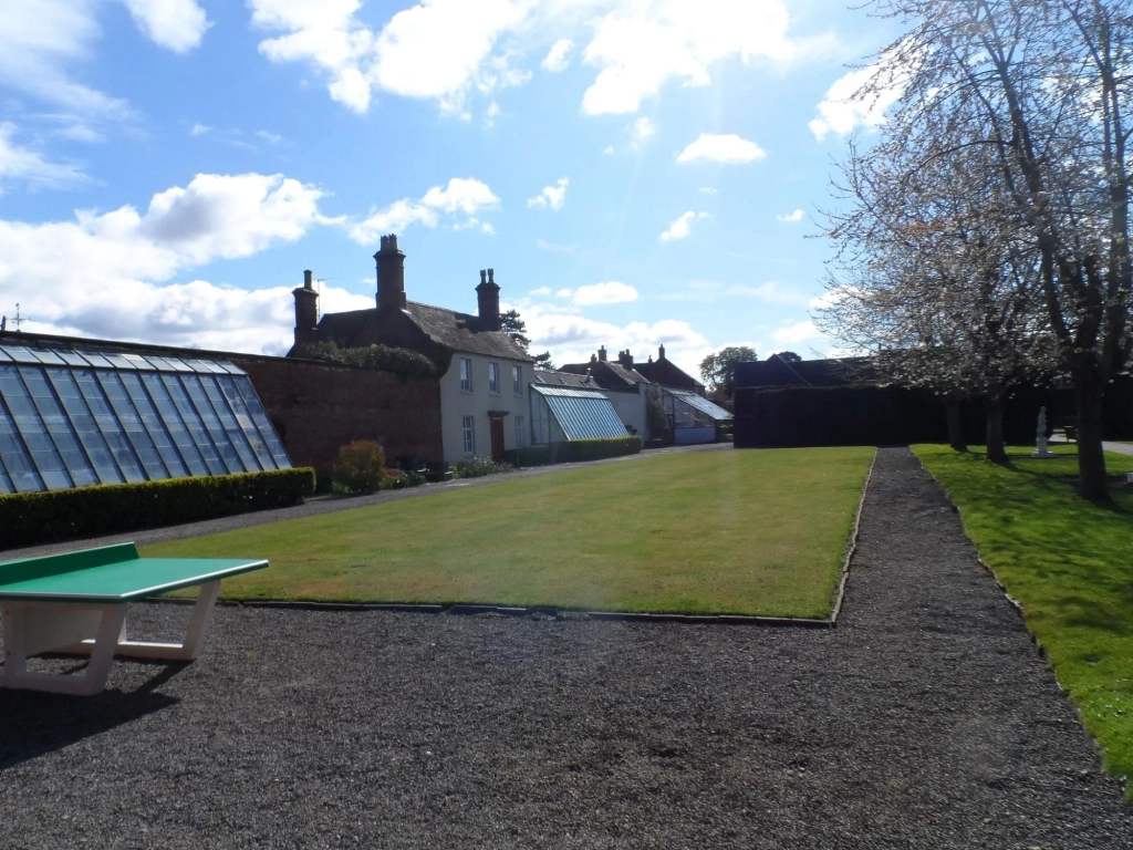 Manicured lawn bowling green flanked by Victorian glasshouses and a cream-painted cottage under a partly cloudy sky.