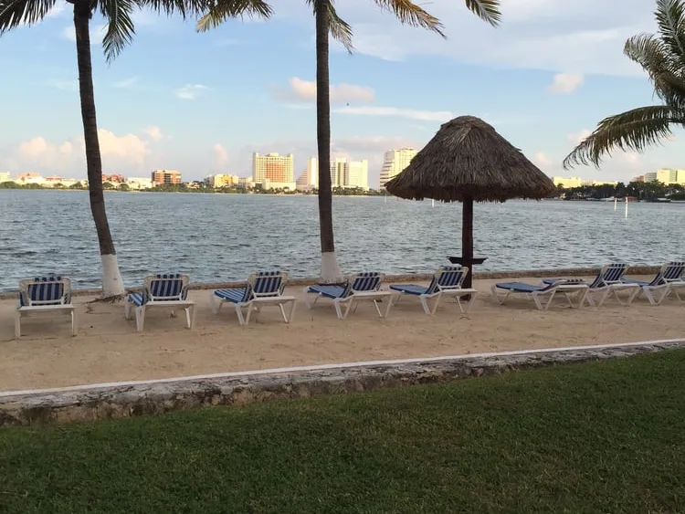 Blue and white lounge chairs on a sandy beach with palm trees, a palapa umbrella, and a city skyline across the water.