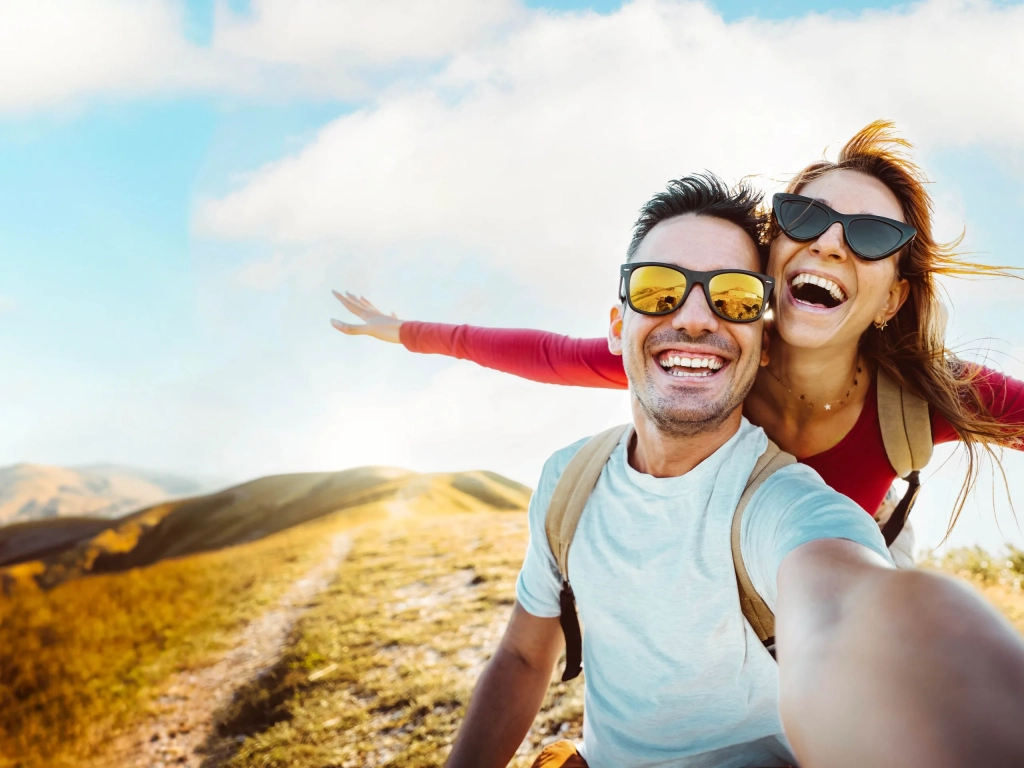 Two smiling hikers wearing sunglasses take a selfie on a sunny mountain trail with rolling hills behind them.