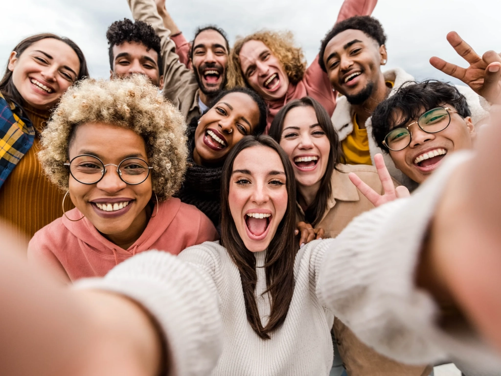 A diverse group of young adults laughing and taking a group selfie outdoors.